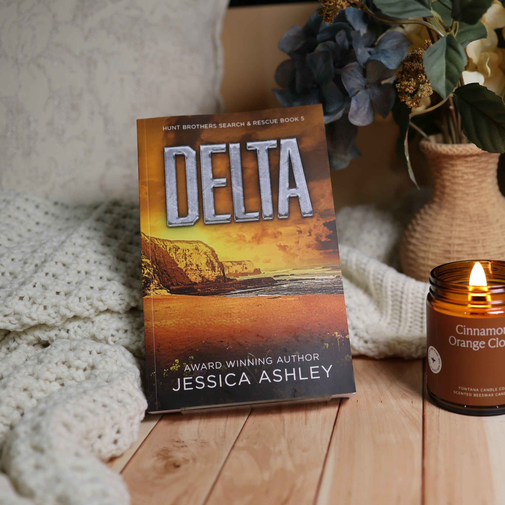 The book 'Delta' by Jessica Ashley is displayed upright on a wooden surface next to a lit candle in a cozy home setting with a knitted blanket and dried flowers.