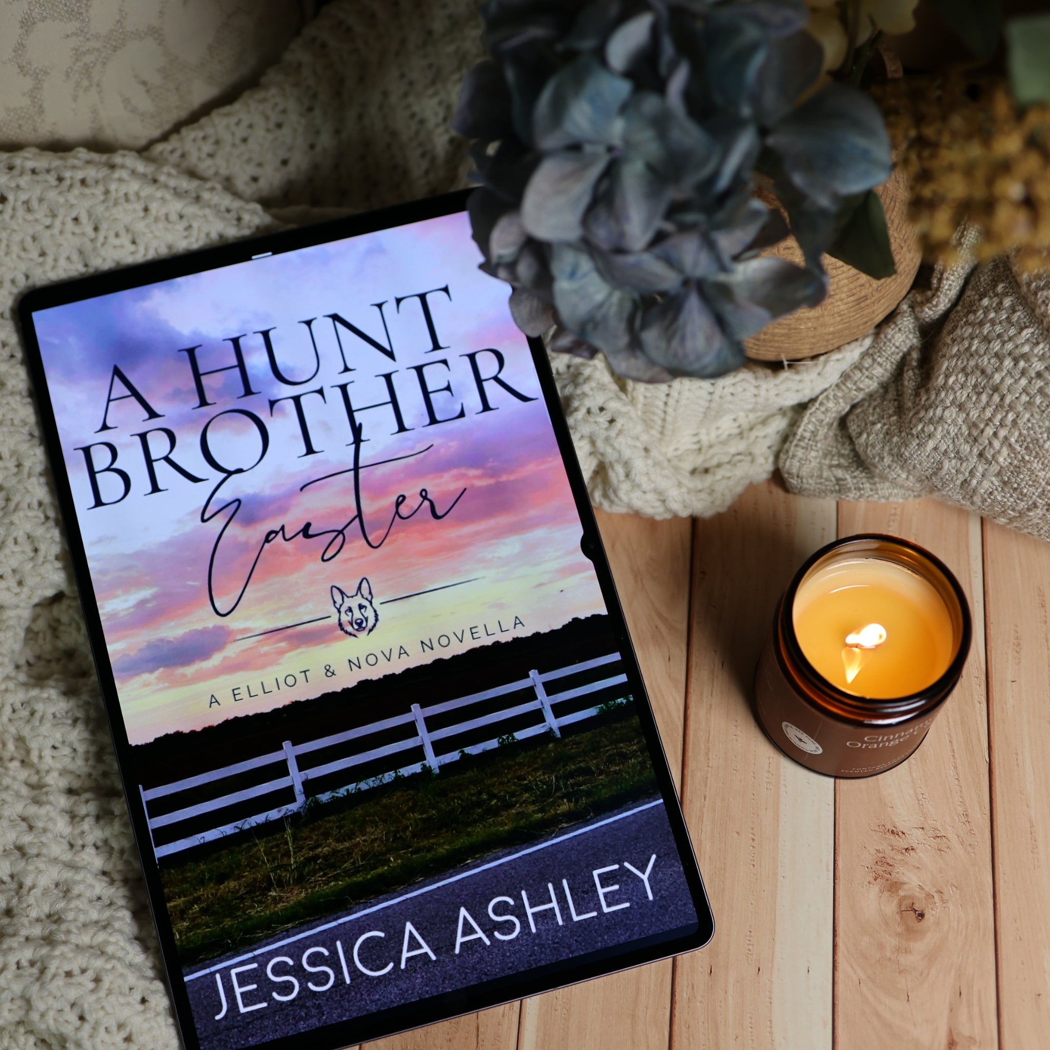 A tablet lying on a wooden surface displays the book cover for 'A Hunt Brother Easter' by Jessica Ashley. The scene is styled with a knitted blanket, a blue hydrangea, and a lit amber candle.