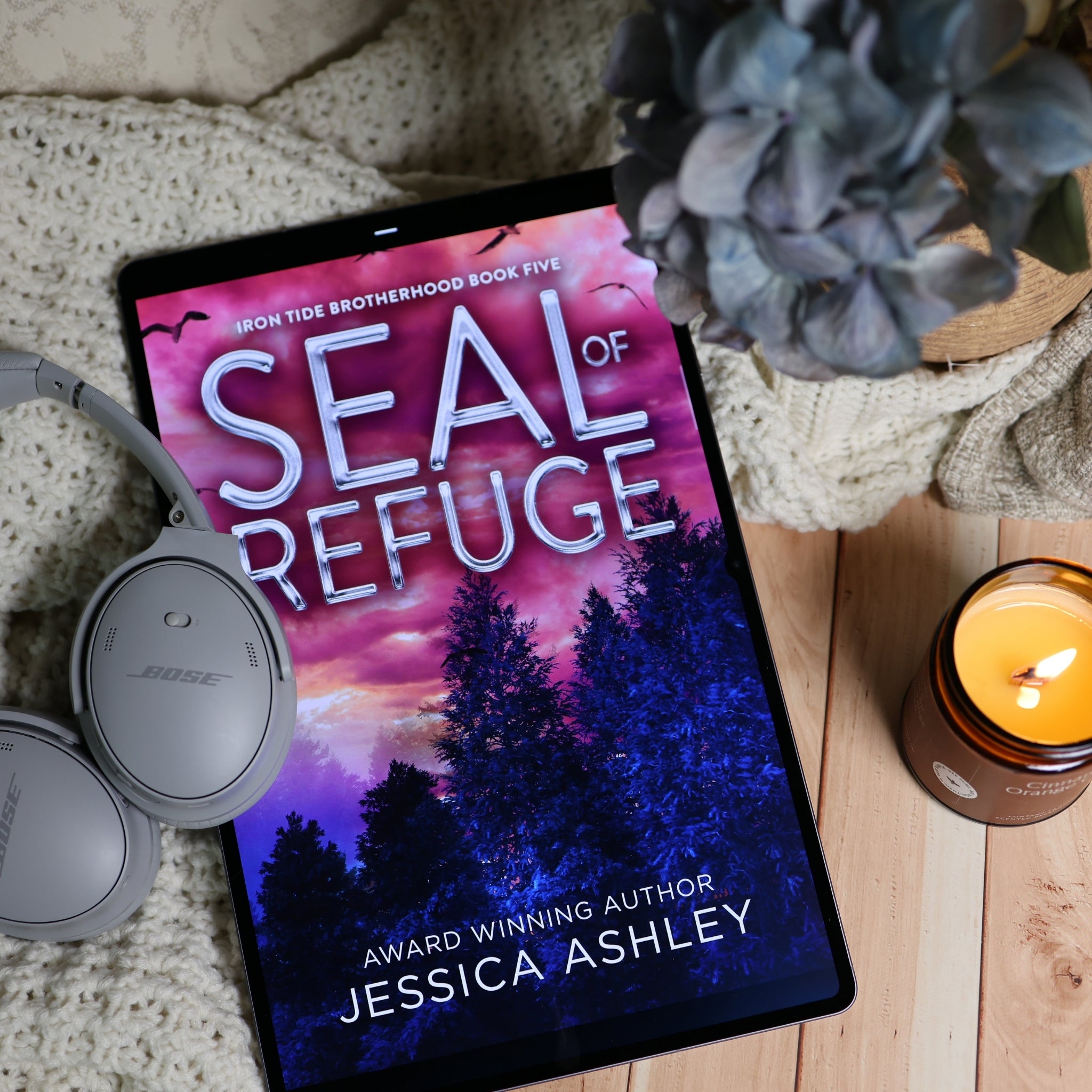 A tablet lying on a wooden surface with a cozy knitted blanket, gray Bose headphones, a lit candle, and a blue flower. The tablet screen displays the audiobook cover for 'Seal of Refuge' by Jessica Ashley.