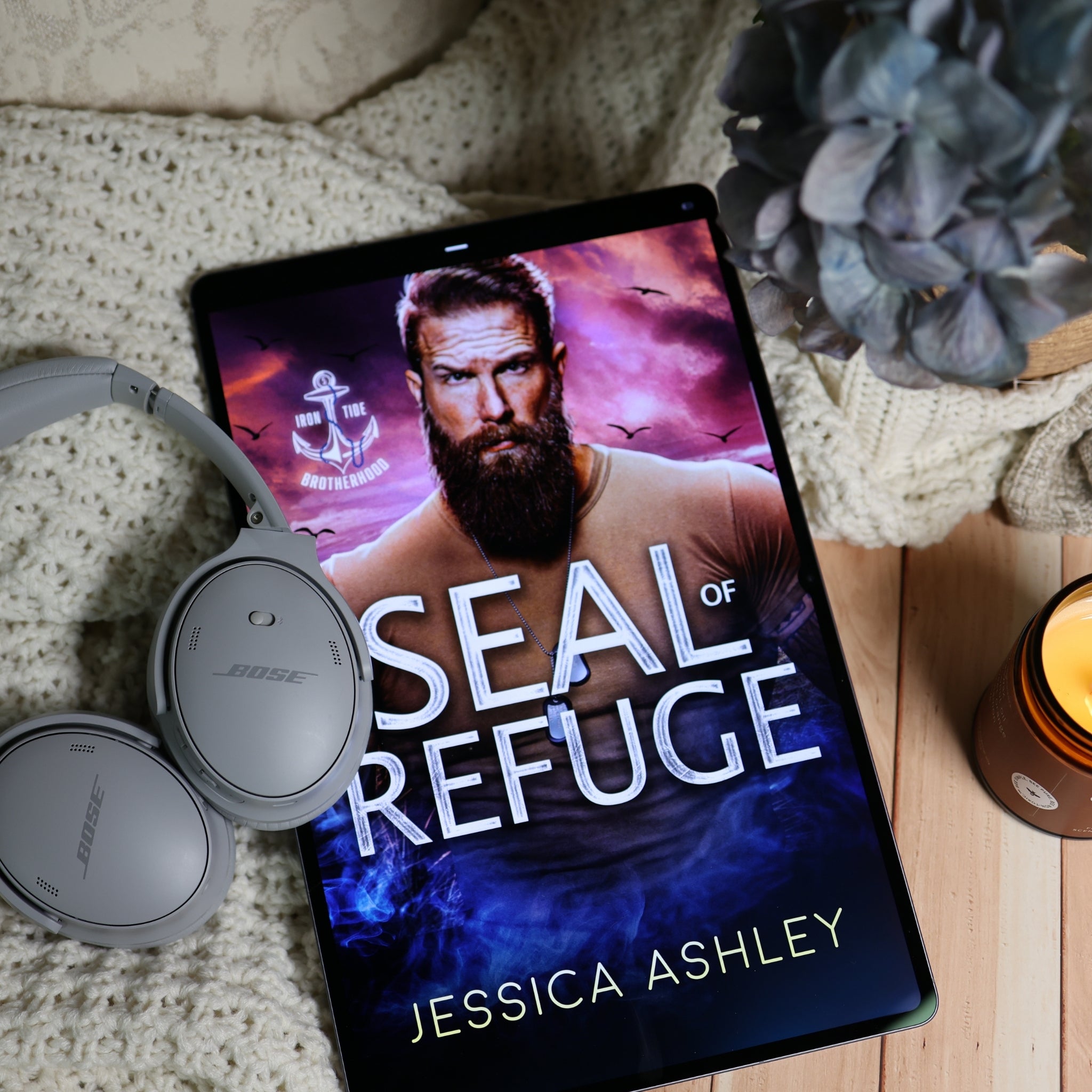 A tablet lying on a cream knit blanket with gray Bose headphones, a blue flower, and a lit candle nearby. The tablet screen displays an audiobook cover for "Seal of Refuge" by Jessica Ashley.