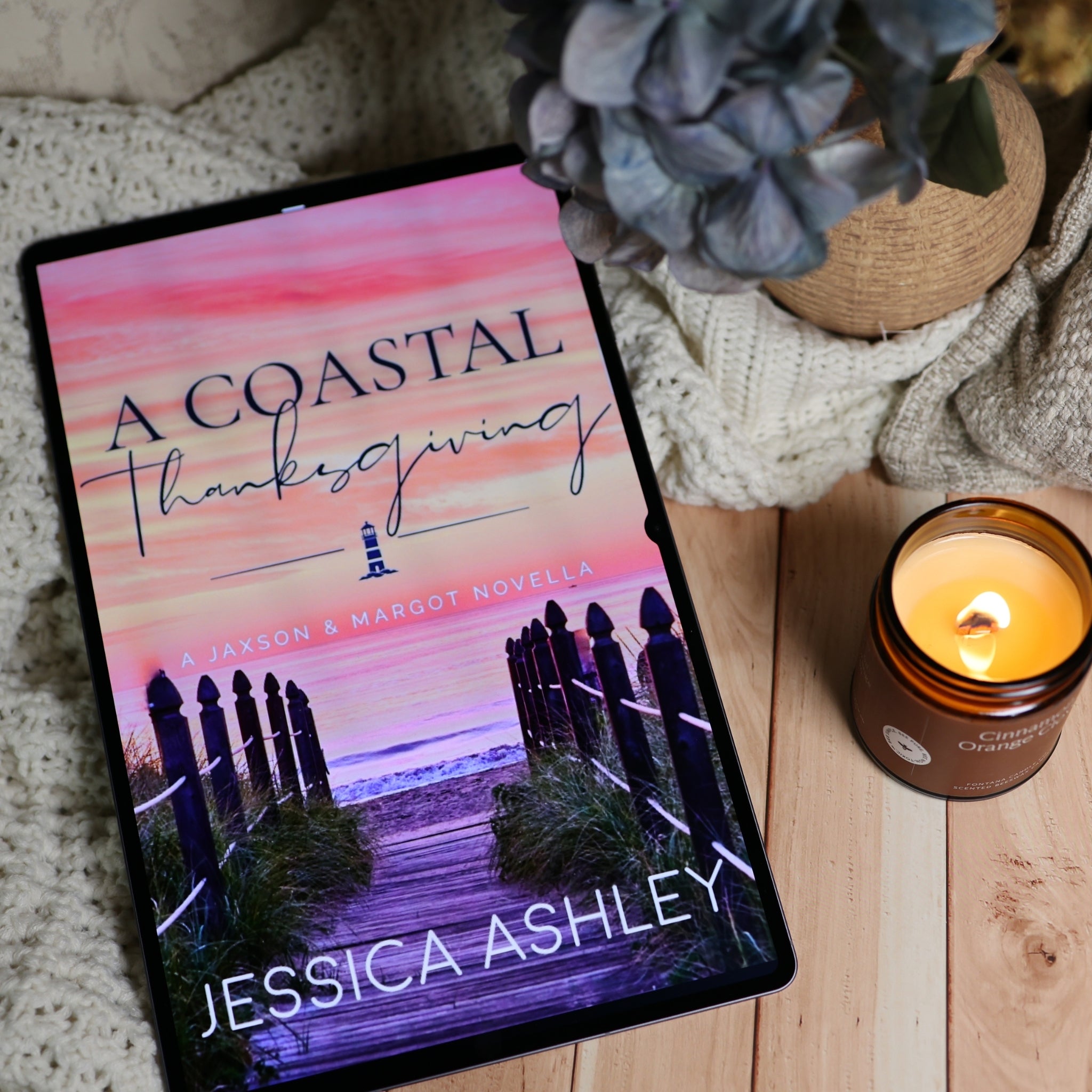 A book titled "A Coastal Thanksgiving" by Jessica Ashley stands upright on a wooden surface next to a lit amber glass candle labeled "Cinnamon Orange Clove." The scene includes a cozy knit blanket, flowers in a vase, and a neutral indoor background.