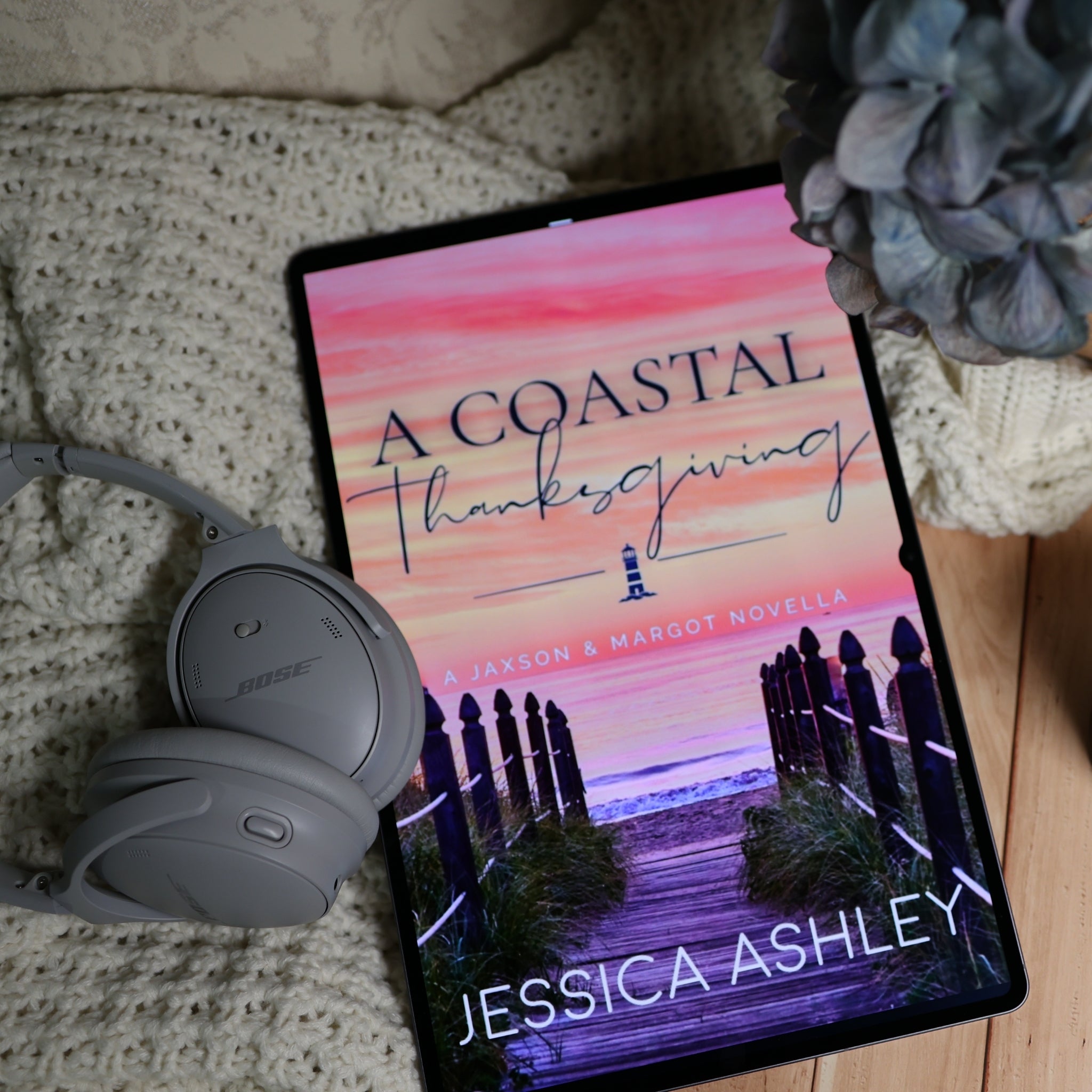 A tablet displaying the book cover of 'A Coastal Thanksgiving' by Jessica Ashley, styled on a beige knit blanket with gray Bose headphones and a blue hydrangea flower.