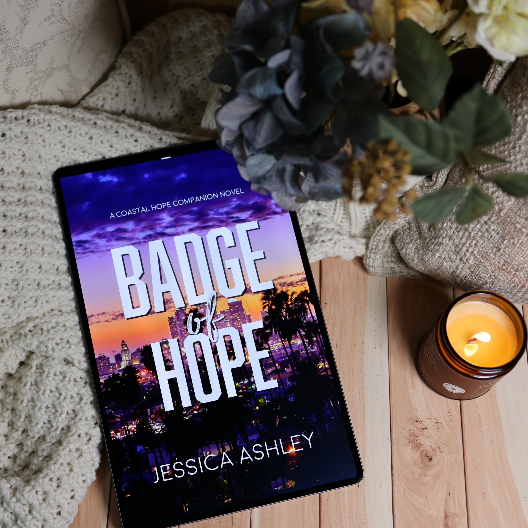 A tablet lying on a wooden surface, displaying the book cover for 'Badge of Hope' by Jessica Ashley. The scene is styled with a cozy knit blanket, dried flowers, and a lit candle.