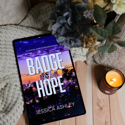A tablet lying on a wooden surface, displaying the book cover for 'Badge of Hope' by Jessica Ashley. The scene is styled with a cozy knit blanket, dried flowers, and a lit candle.