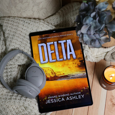 A tablet lying on a cozy knit blanket with gray Bose headphones, a lit candle, and a blue flower, displaying the book cover for 'Delta' by Jessica Ashley.