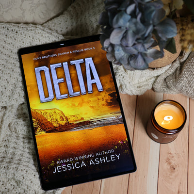 A tablet displaying the book cover of 'Delta' by Jessica Ashley, styled on a wooden surface with a knitted blanket, a lit candle, and a potted plant.