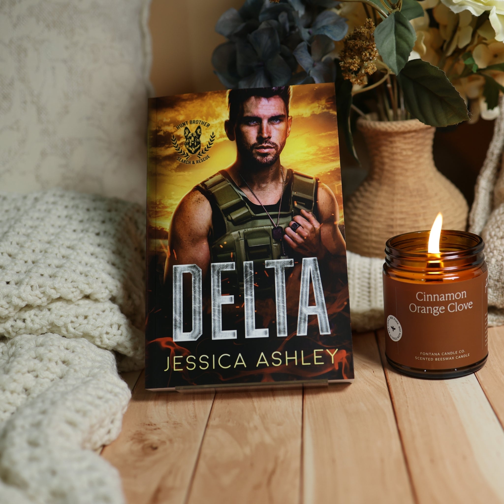 A book titled 'Delta' by Jessica Ashley stands upright on a wooden surface next to a lit amber glass candle labeled 'Cinnamon Orange Glove.' The scene includes a cozy knit blanket, dried flowers in a woven vase, and a warm indoor setting.