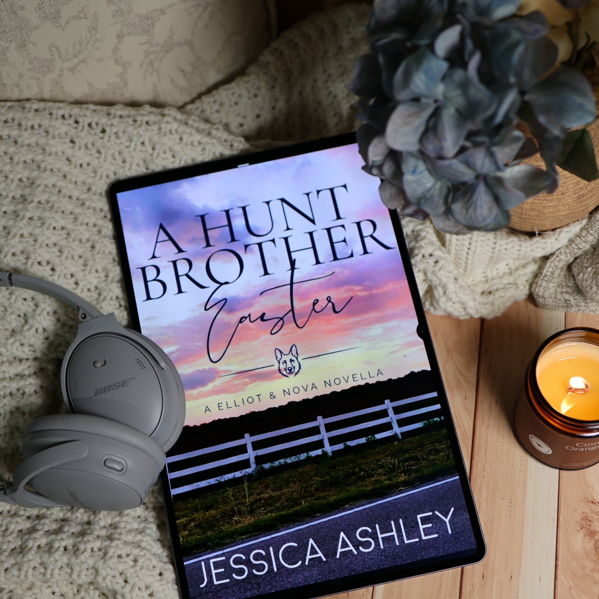 A tablet displaying the book cover of 'A Hunt Brother Easter' by Jessica Ashley, styled in a cozy bedroom setting with a knit blanket, gray Bose headphones, a blue hydrangea, and a burning candle.