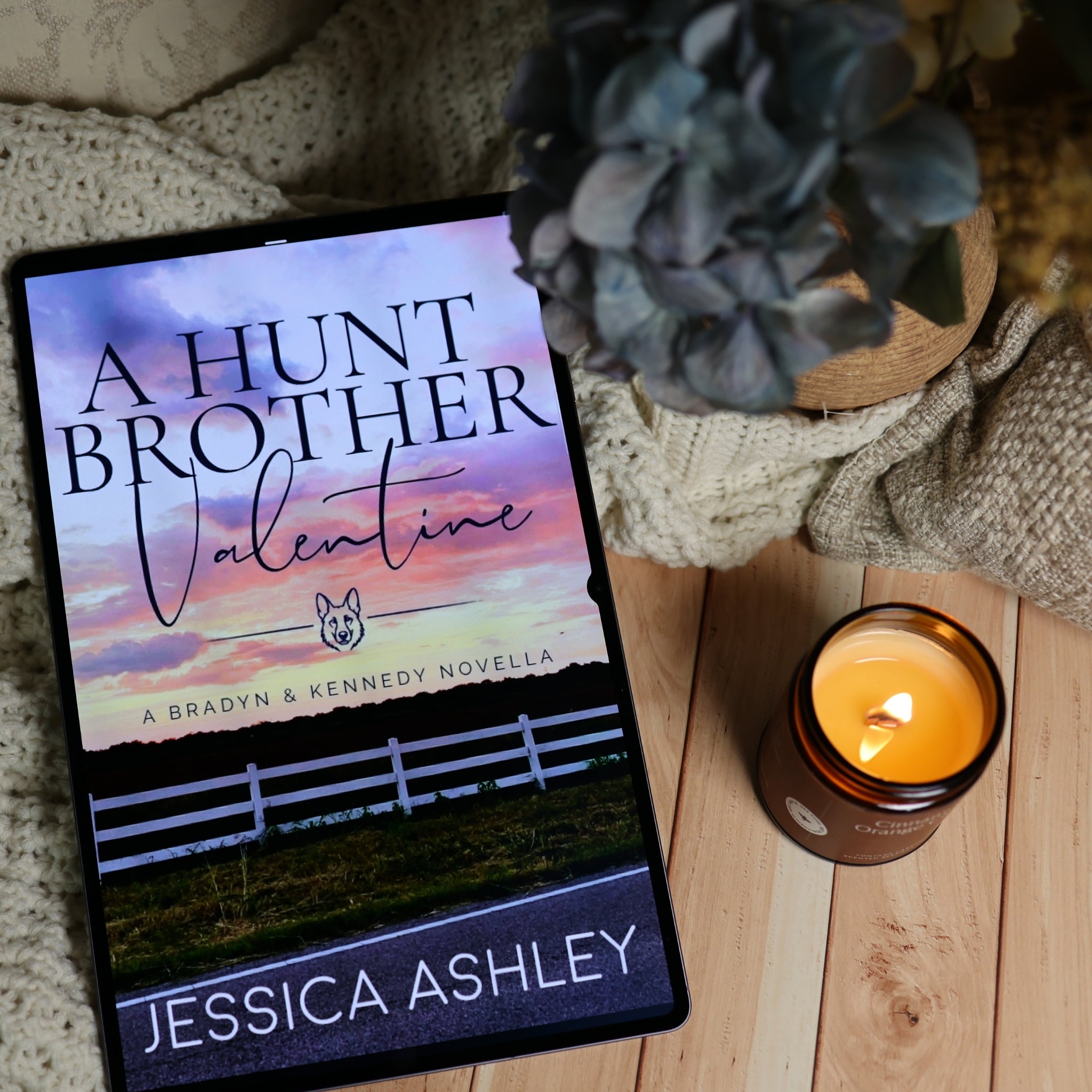 A tablet displaying the book cover of 'A Hunt Brother Valentine' by Jessica Ashley, styled on a wooden surface with a lit candle and a knitted blanket nearby.