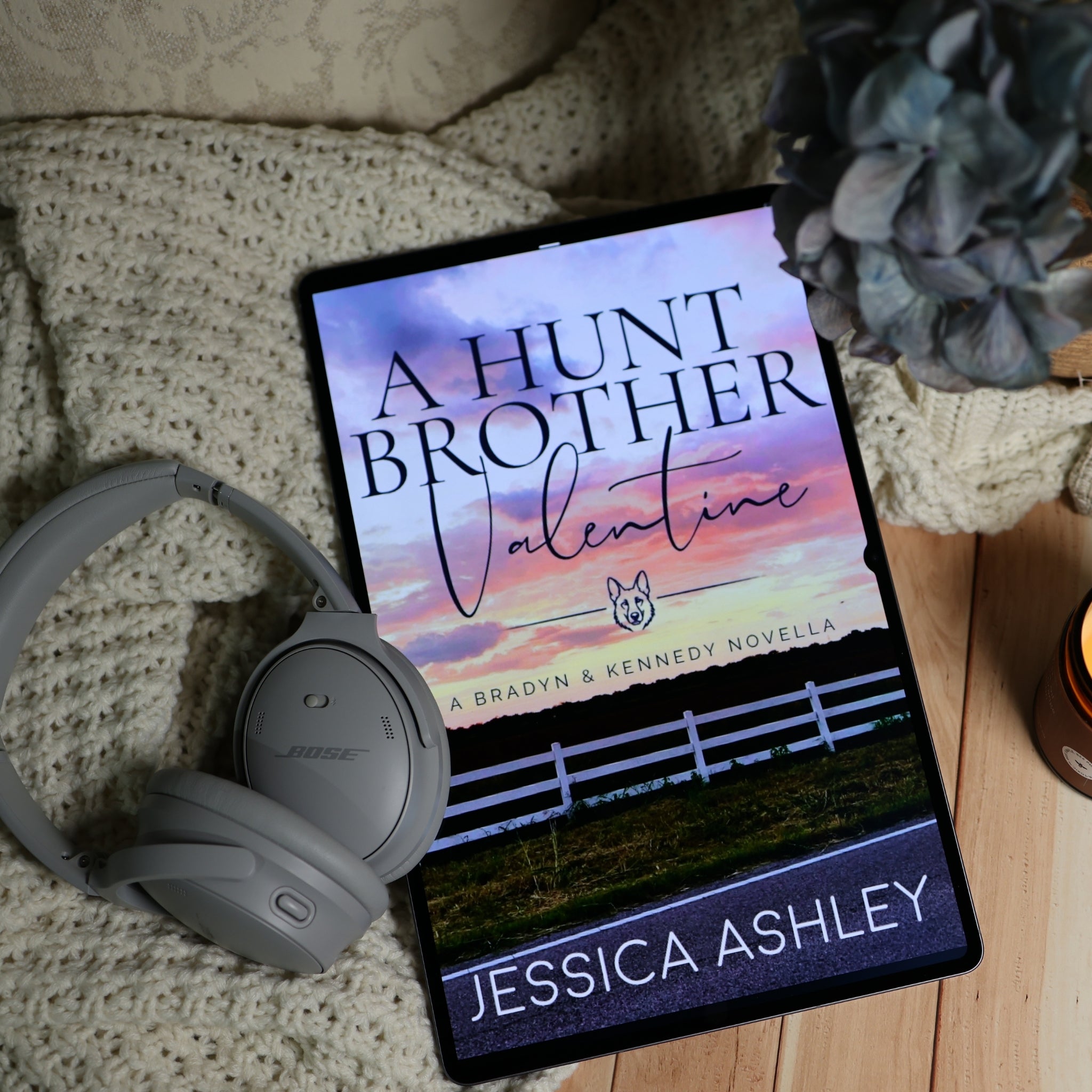 A tablet displaying the book cover of 'A Hunt Brother Valentine' by Jessica Ashley, styled on a wooden surface with a beige knit blanket, gray Bose headphones, and a blue flower.