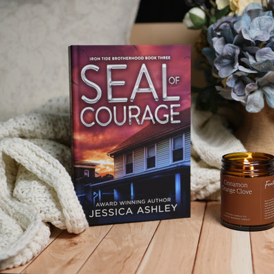 Book cover for 'Seal of Courage' by Jessica Ashley, styled on a wooden surface next to a lit candle and a cream knit blanket, with blue flowers in the background.