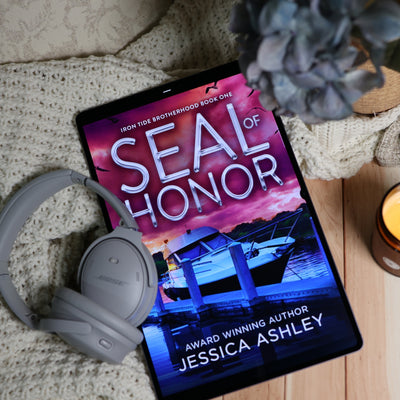 A tablet lying on a beige knit blanket with gray Bose headphones resting beside it. The screen displays the book cover for 'Seal of Honor' by Jessica Ashley, with a pink and purple sunset sky, a boat, and blue water.