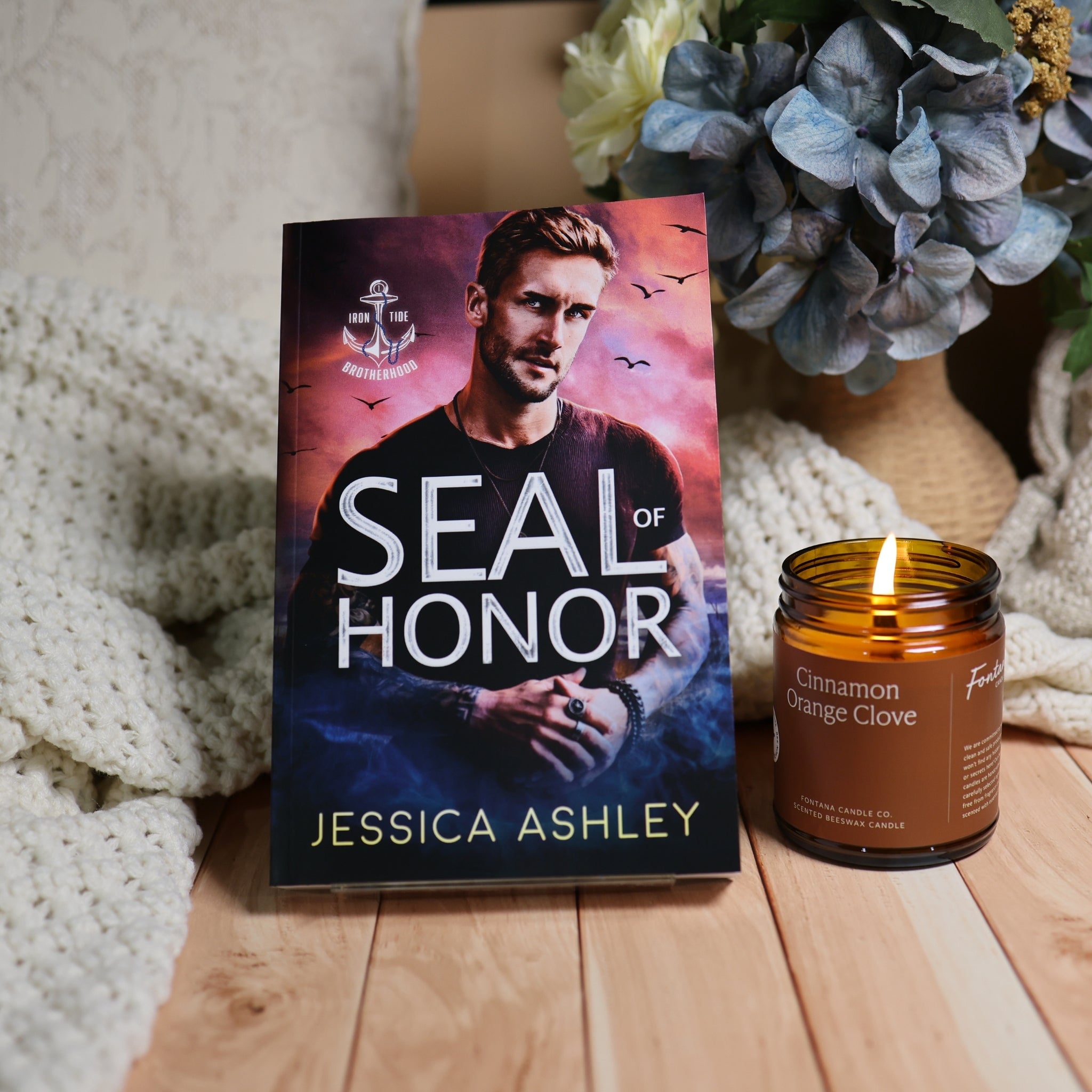 A book titled 'Seal of Honor' by Jessica Ashley stands upright on a wooden surface next to a lit amber candle jar. The scene is styled with a cream knit blanket and blue hydrangeas in the background.