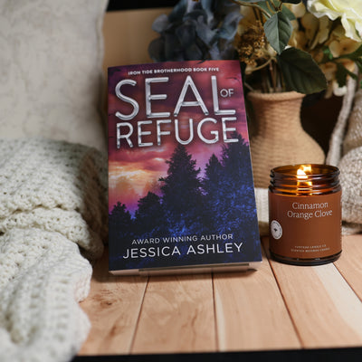 A book titled "Seal of Refuge" by Jessica Ashley stands upright on a wooden surface next to a lit amber glass candle labeled "Cinnamon Orange Clove." The scene is styled with a cream knit blanket, dried flowers in a woven vase, and a neutral background.