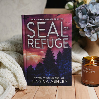 The book 'Seal of Refuge' by Jessica Ashley stands upright on a wooden surface, styled with a cream knit blanket on the left and a lit 'Cinnamon Orange Cleve' candle on the right, against a background of dried flowers.