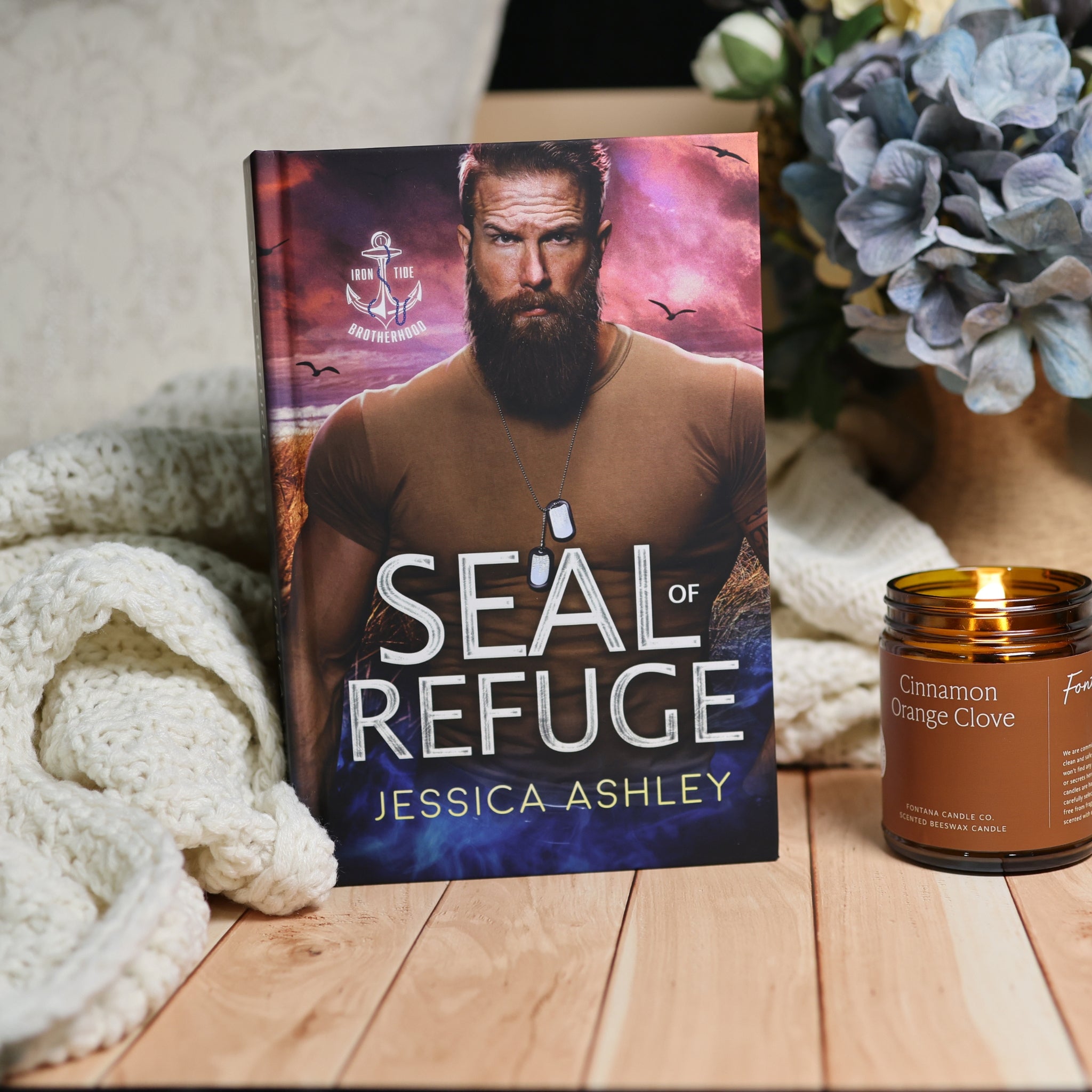 A book titled "Seal of Refuge" by Jessica Ashley stands upright on a wooden surface, styled with a cream knit blanket on the left and a lit amber candle labeled "Cinnamon Orange Clove" on the right. The cover art shows a bearded man against a dramatic purple and pink sky.