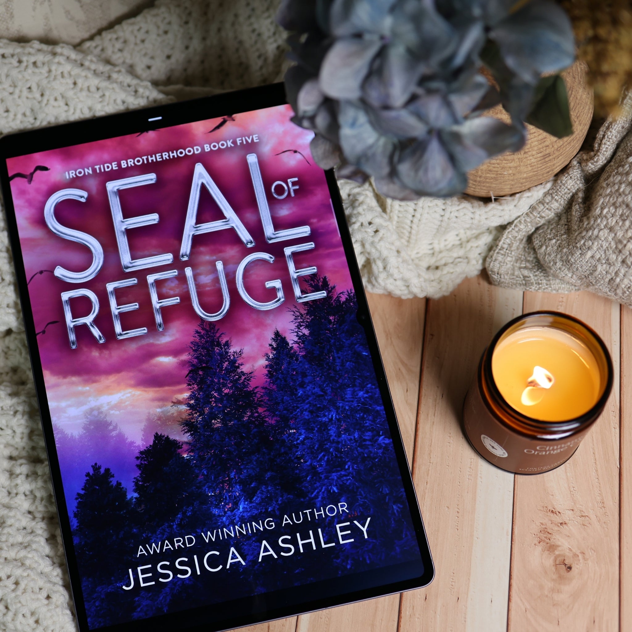 A tablet displaying the book cover of 'Seal of Refuge' by Jessica Ashley, styled on a wooden surface with a knitted blanket, a lit amber candle, and a dried flower.