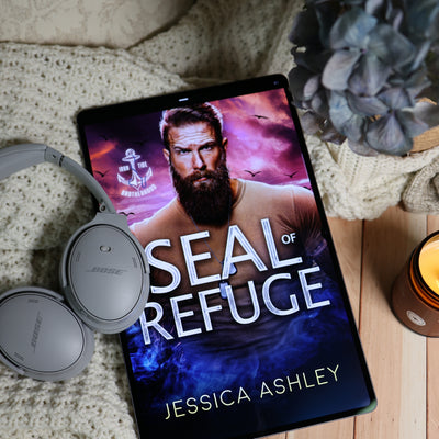 A tablet lying on a cream knit blanket with gray Bose headphones, a blue flower, and a lit candle nearby. The tablet screen displays an audiobook cover for "Seal of Refuge" by Jessica Ashley.