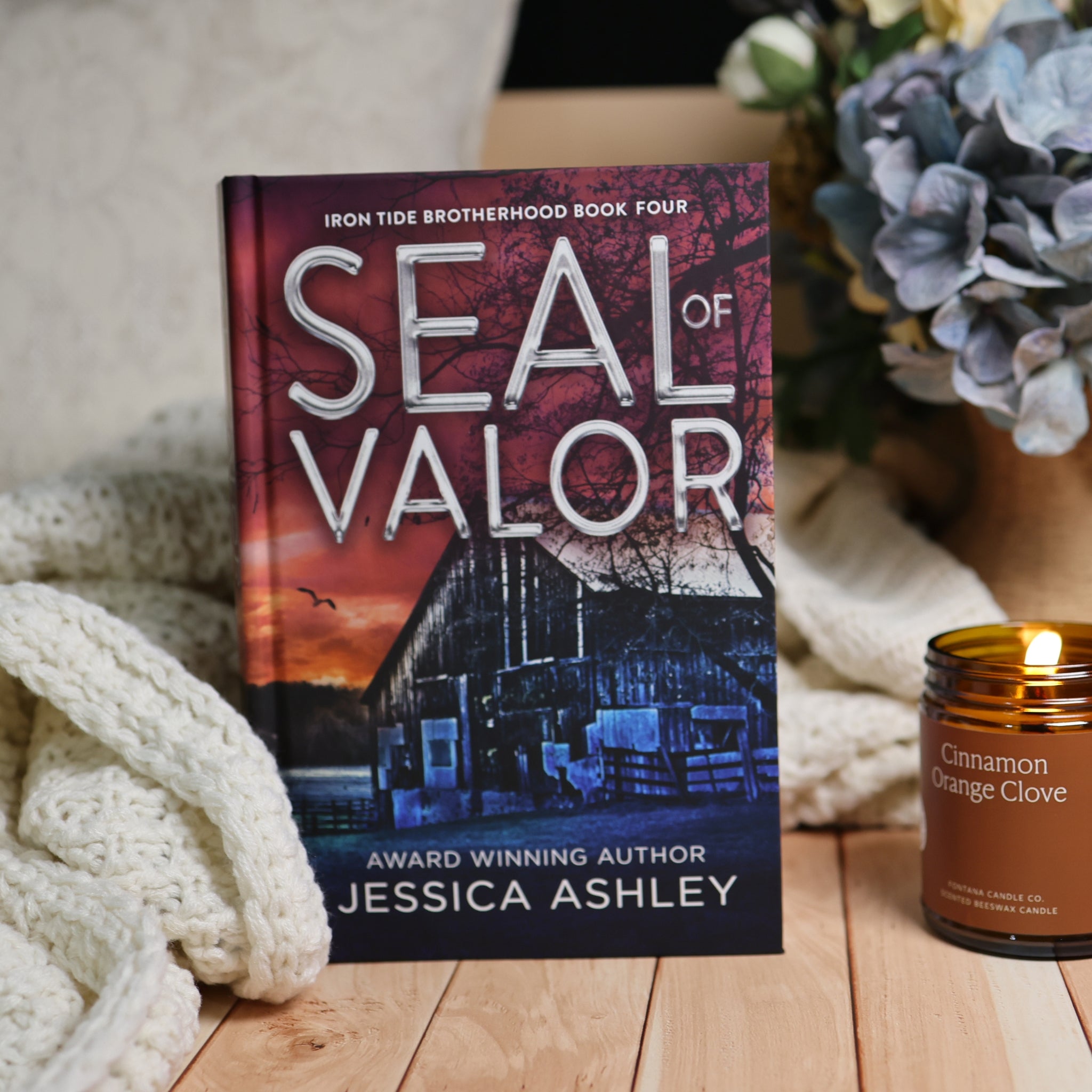 Book cover for 'Seal of Valor' by Jessica Ashley, styled on a wooden surface with a cream knit blanket, a lit candle, and blue flowers in the background.