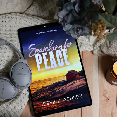 A tablet lying on a wooden surface, displaying the book cover for "Searching for Peace" by Jessica Ashley. The scene is styled with a knit blanket, gray Bose headphones, dried flowers, and a candle, creating a cozy reading atmosphere.