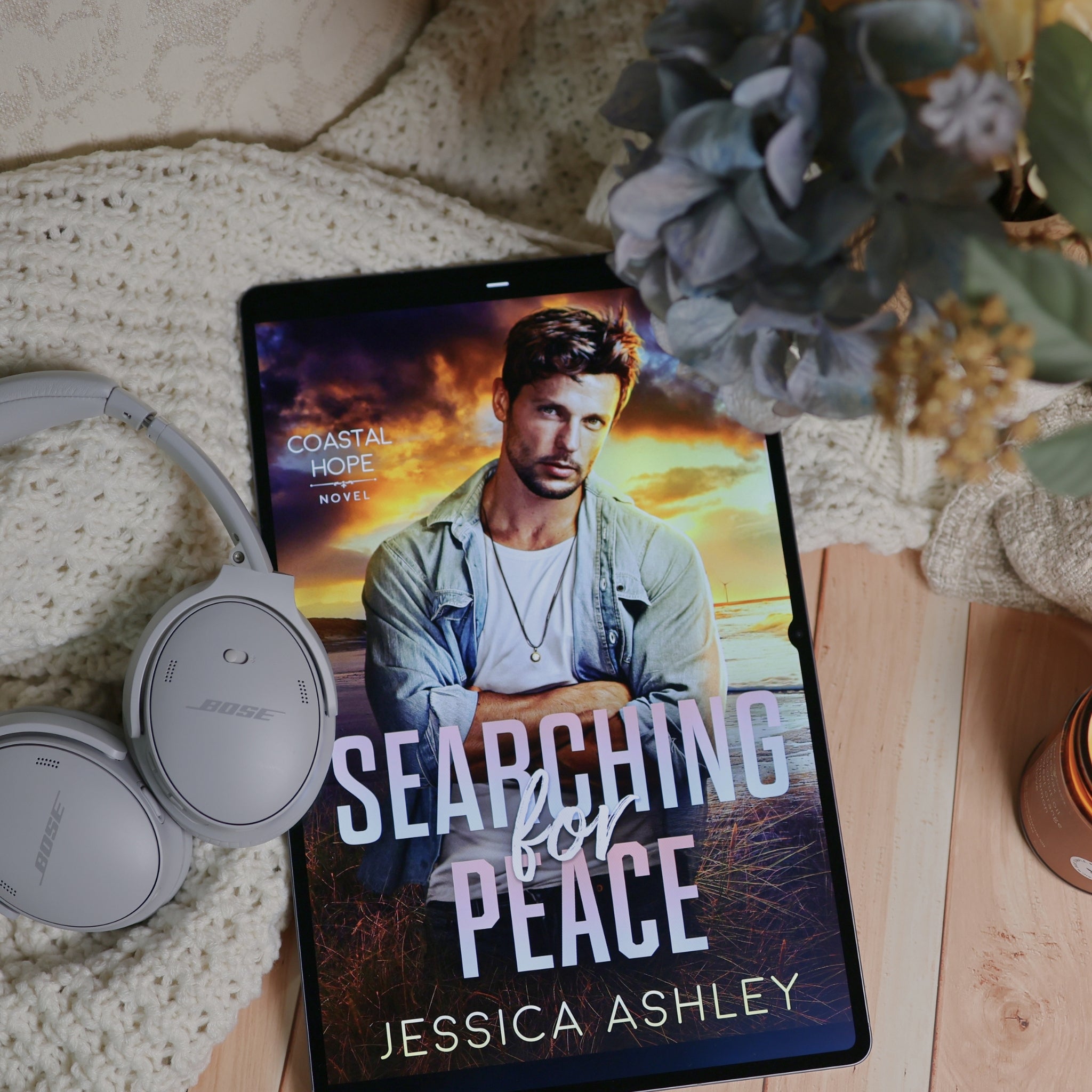 A tablet displaying the book cover of 'Searching for Peace' by Jessica Ashley, styled in a cozy flat-lay with white headphones, a knit blanket, and dried flowers on a wooden surface.