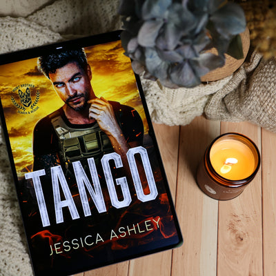 A tablet displaying the book cover of 'Tango' by Jessica Ashley, styled in a cozy flat-lay setting with a lit candle, a knit blanket, and a potted plant on a light wood surface.